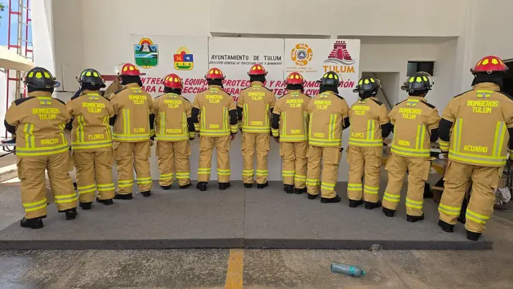 Firefighters in yellow and tan uniforms and helmets stand in a line facing away from the camera inside a building with municipal logos on the wall behind them