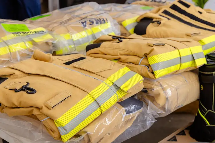Tan firefighting jackets with reflective yellow safety stripes are displayed on a table, wrapped in protective plastic