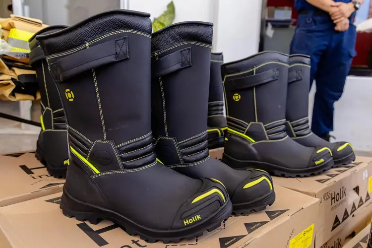 Black and yellow firefighting boots displayed on a cardboard box during an indoor presentation, with additional boots visible behind and a person in blue unifor