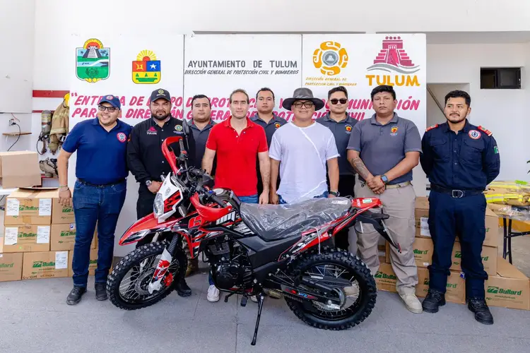Ten firefighters stand beside a red and black motorcycle in an indoor facility with municipal logos and banners on the wall behind them