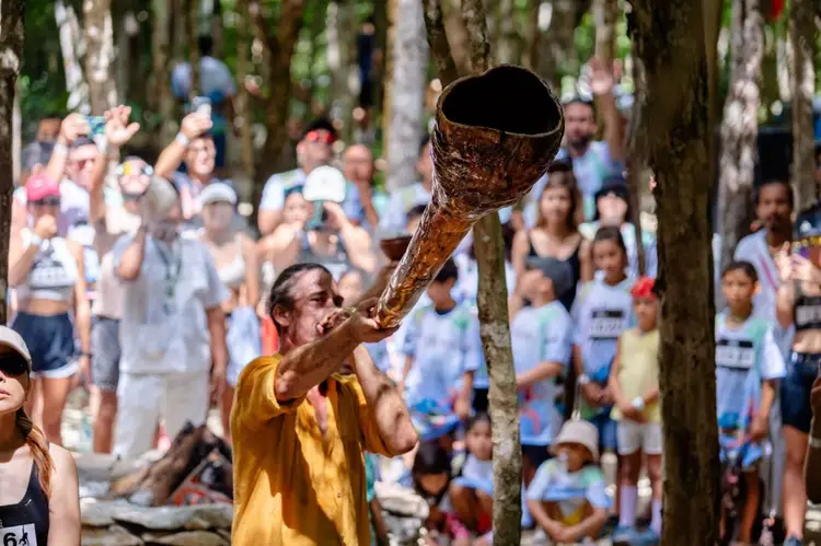 Man in yellow shirt holds up a large woven basket while surrounded by spectators in a forested area