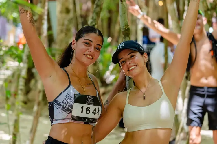 Two female runners with arms raised celebrate under a canopy of hanging vines in a jungle setting during a trail race