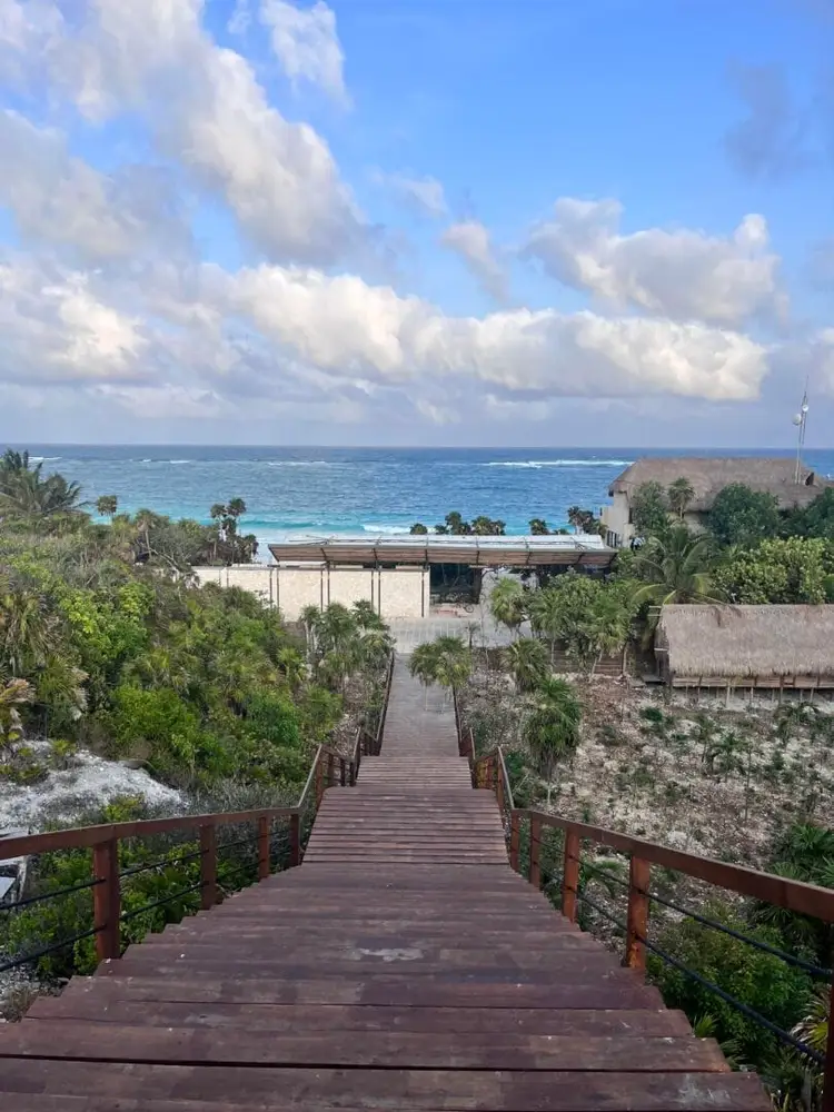 Wooden boardwalk descends toward a beachfront structure and turquoise ocean, surrounded by tropical vegetation and thatched-roof buildings Wooden boardwalk descends toward a beachfront structure and turquoise ocean, surrounded by tropical vegetation and thatched-roof buildings