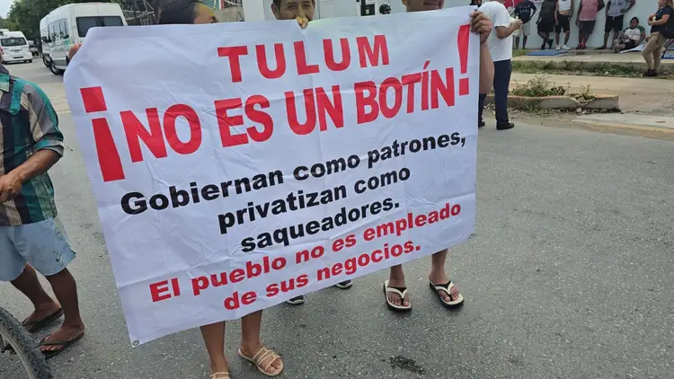Protesters hold a white banner with red and black Spanish text during a street demonstration, with vehicles and bystanders visible in the background