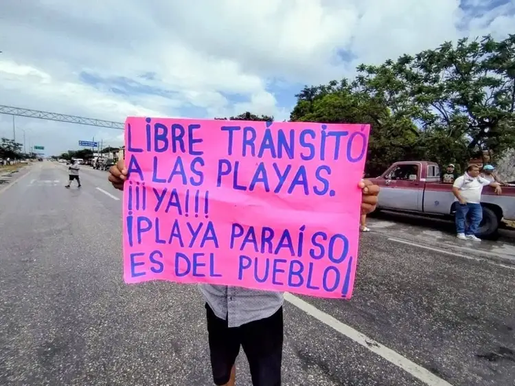 A protester holds a bright pink sign with Spanish text while standing on a roadway, with other people and vehicles visible behind them in a residential area