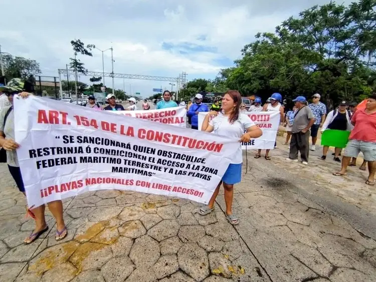 Protesters gather on a street holding a large banner with Spanish text about constitutional rights and beach access, with others standing behind them under clou