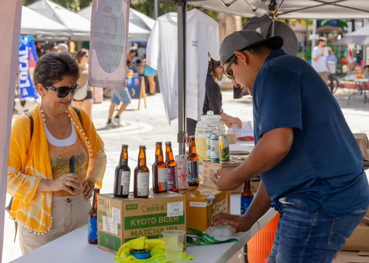 A vendor serves a customer at an outdoor market beer stand displaying bottles under a white tent canopy A vendor serves a customer at an outdoor market beer stand displaying bottles under a white tent canopy