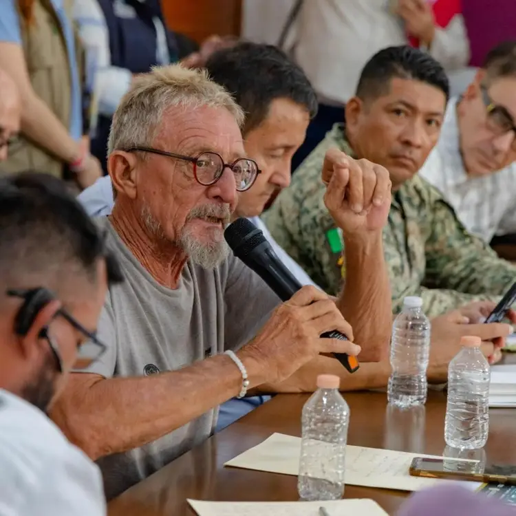 Man with glasses and gray beard speaks into microphone at a table during what appears to be a public meeting or forum