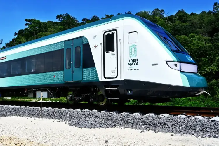 A modern passenger train with white and teal livery sits on gravel alongside dense green forest under clear blue sky A modern passenger train with white and teal livery sits on gravel alongside dense green forest under clear blue sky