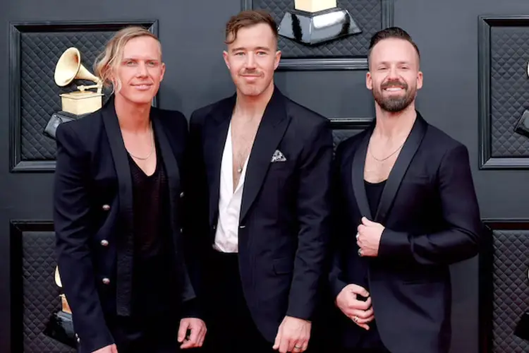 Three men in dark blazers stand together in front of a gray textured wall with gold gramophone decorations Three men in dark blazers stand together in front of a gray textured wall with gold gramophone decorations