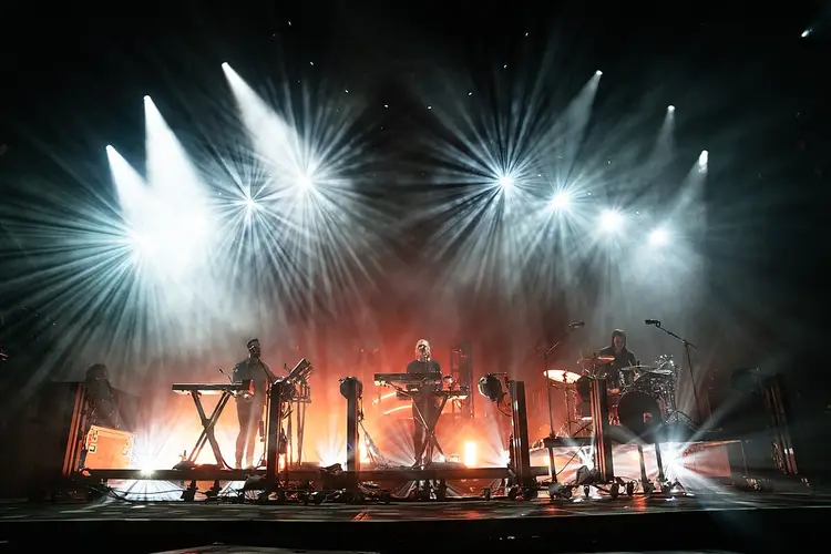A band performs on stage with bright white overhead lights and warm orange backlighting, with multiple musicians playing keyboards and drums A band performs on stage with bright white overhead lights and warm orange backlighting, with multiple musicians playing keyboards and drums