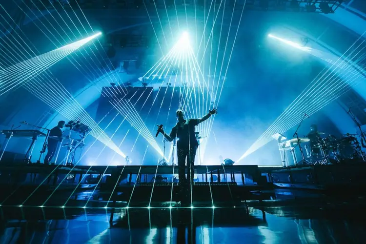 Performer on stage with arm raised, silhouetted against dramatic blue lighting and laser beams, with band members and instruments visible on either side Performer on stage with arm raised, silhouetted against dramatic blue lighting and laser beams, with band members and instruments visible on either side