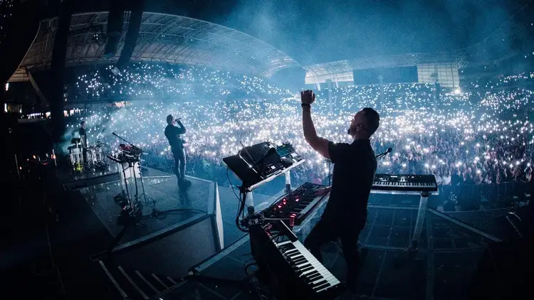 Musicians perform on stage with electronic instruments before a large crowd in an arena lit by thousands of phone lights Musicians perform on stage with electronic instruments before a large crowd in an arena lit by thousands of phone lights