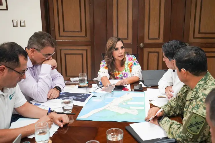 Five people sit around a wooden table reviewing a map during an indoor meeting, with water glasses and documents visible