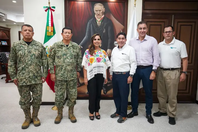 Six people stand in a line in an official interior setting with a Mexican flag and portrait on the wall behind them, including two in military uniforms and four