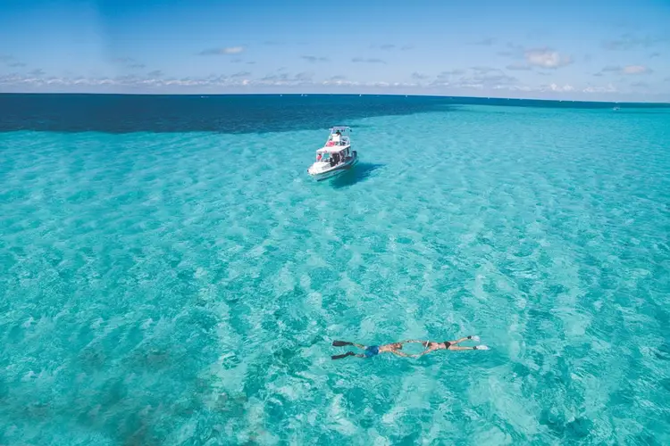 A person snorkels in shallow turquoise water while a white boat floats nearby under a clear blue sky