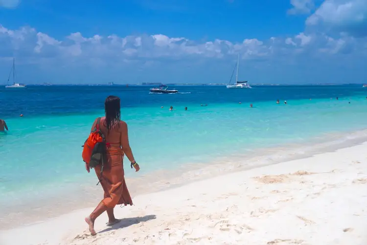 Woman in orange dress carrying a red float walks along a sandy beach toward turquoise water with sailboats anchored offshore