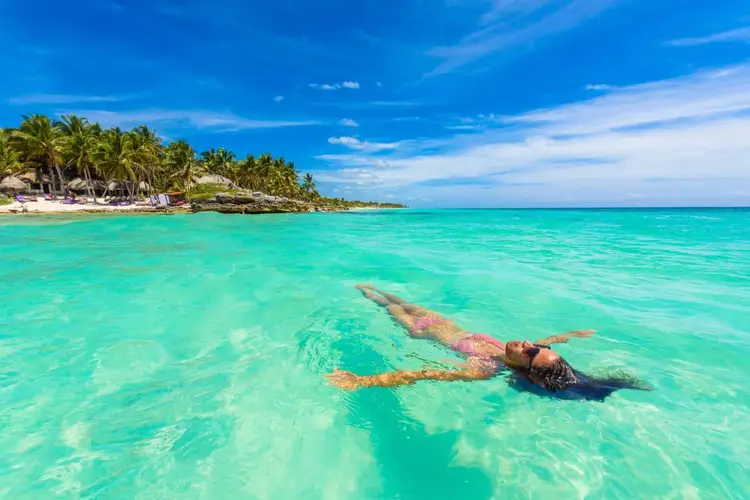 Person snorkeling in clear turquoise water near a palm-lined beach with white sand
