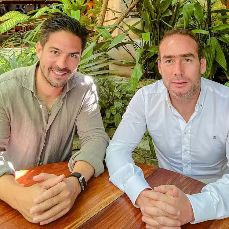Two men sit at a wooden table surrounded by potted plants, one holding a small orange object in his hand Two men sit at a wooden table surrounded by potted plants, one holding a small orange object in his hand