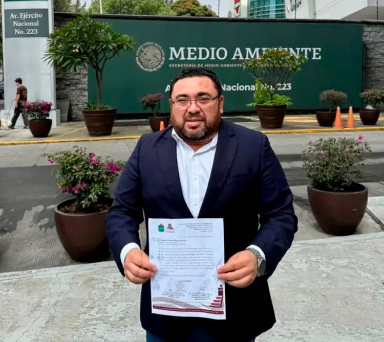 Man in dark blazer and glasses holds a document while standing in front of a green government building with potted flowers