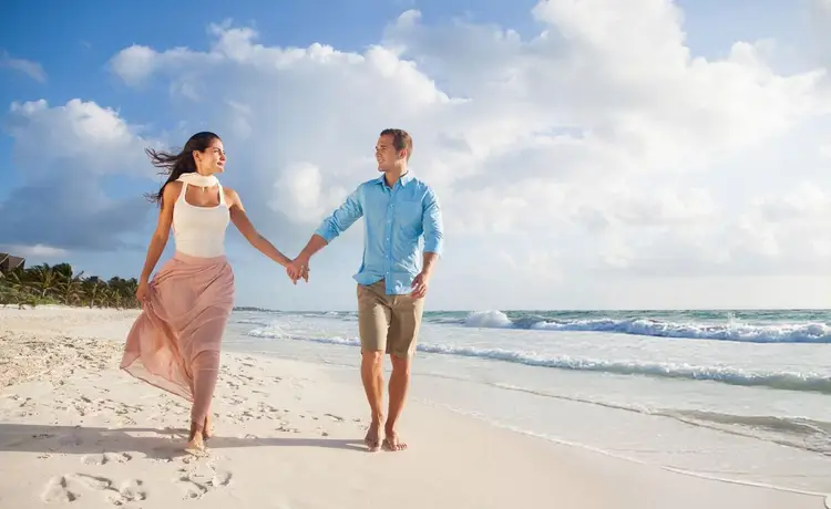 Two people hold hands while walking along a sandy beach with waves and clouds in the background Two people hold hands while walking along a sandy beach with waves and clouds in the background