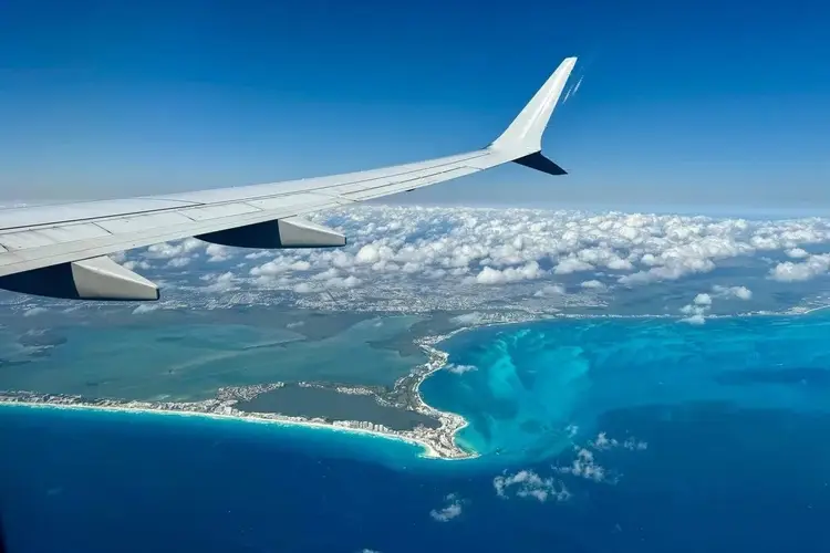 Airplane wing and engine frame a curved coastline with turquoise waters and white sand beaches below a partly cloudy sky