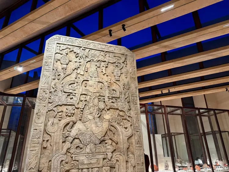 Stone carved relief panel with intricate figural and symbolic designs displayed indoors beneath wooden beams and blue evening sky