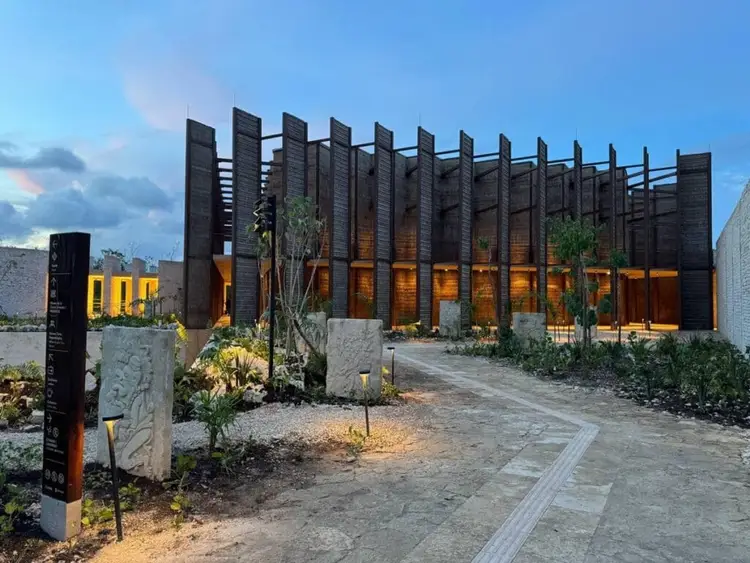 Modern building with dark wooden slatted exterior walls and illuminated orange interior, surrounded by landscaping and a paved pathway at dusk