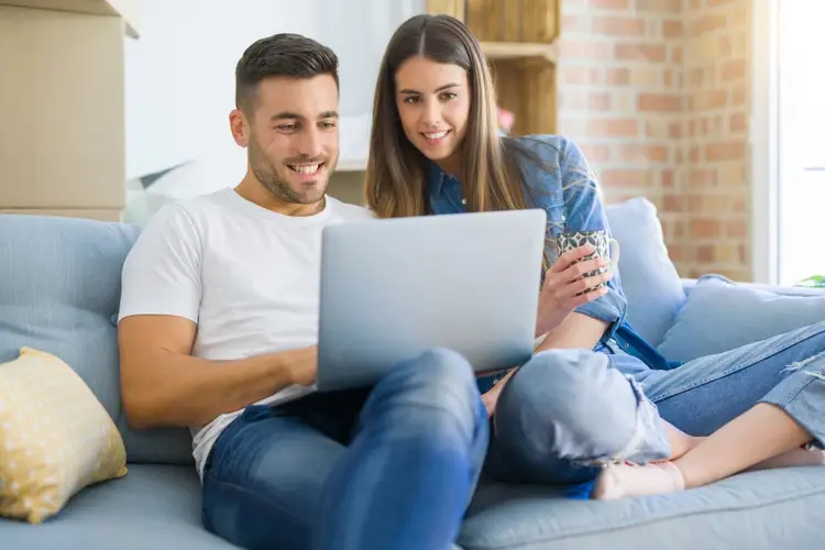 Two people sit on a gray couch looking at a laptop together in a brick and white interior space Two people sit on a gray couch looking at a laptop together in a brick and white interior space