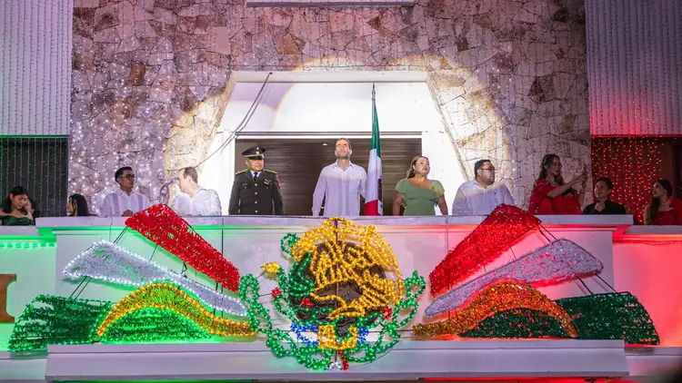 Officials and attendees stand on a balcony above a display featuring the Mexican coat of arms made from colorful beads and flowers