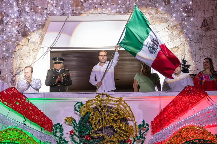 Man in white shirt waves Mexican flag from a balcony decorated with lights while officials and spectators watch below