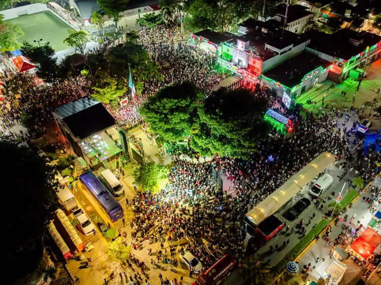 Aerial view of a large nighttime crowd gathered in a town square surrounded by colorful stage lighting and buildings