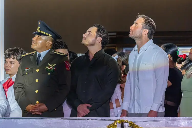 A military officer in uniform stands with two men in civilian clothing and others at what appears to be an indoor public event or gathering