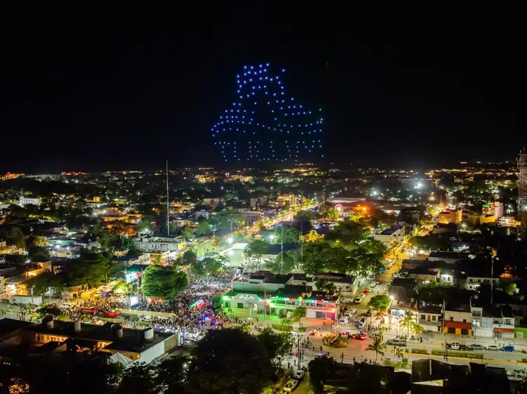 Nighttime aerial view of a town square filled with crowds below blue glowing lights forming a pattern in the night sky above illuminated buildings and streets
