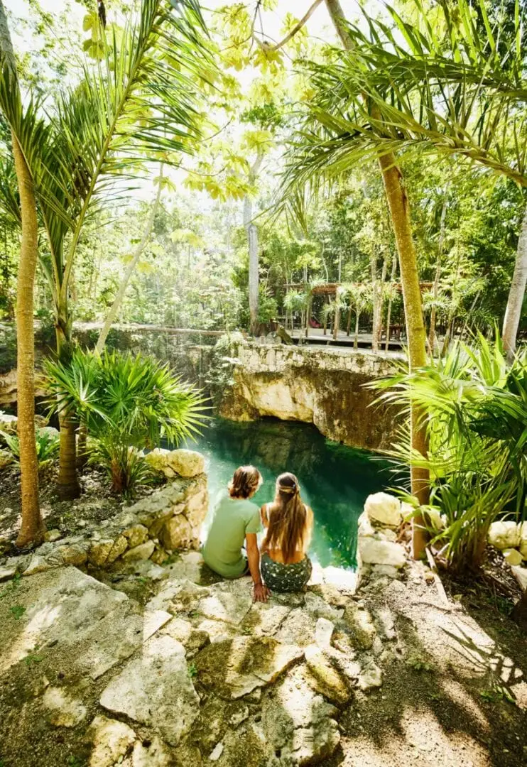 Two visitors sit on rocky ground overlooking a turquoise cenote surrounded by tall palm trees and a wooden bridge in a dense jungle setting