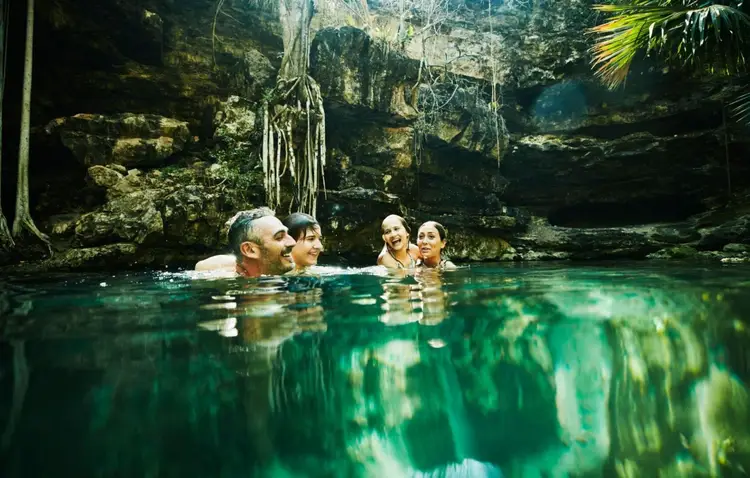 Four people swim in clear turquoise water surrounded by tall trees, vegetation, and rope hanging from cave walls above