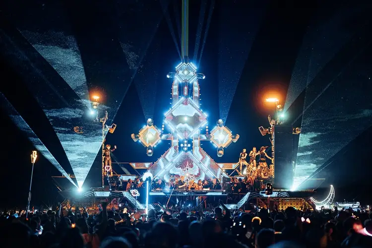 Nighttime festival crowd gathers before an illuminated sculptural stage structure with geometric designs and dramatic lighting effects Nighttime festival crowd gathers before an illuminated sculptural stage structure with geometric designs and dramatic lighting effects