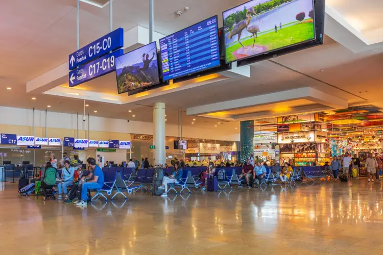 Passengers wait in airport terminal seating area beneath flight information displays and overhead signage
