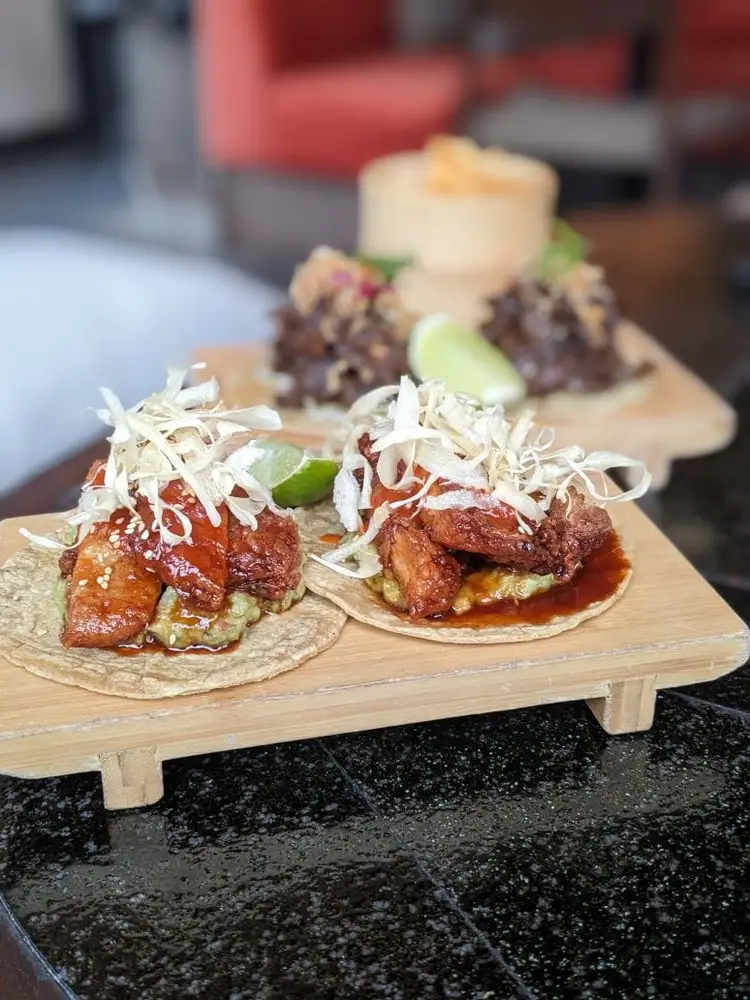 Two tacos with braised meat, shredded cabbage, and lime sit on a wooden board in a restaurant setting