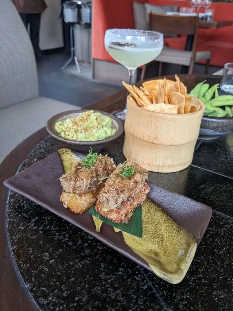 Plated fried appetizers with guacamole dip and tortilla chips sit on a restaurant table with a cocktail in the background
