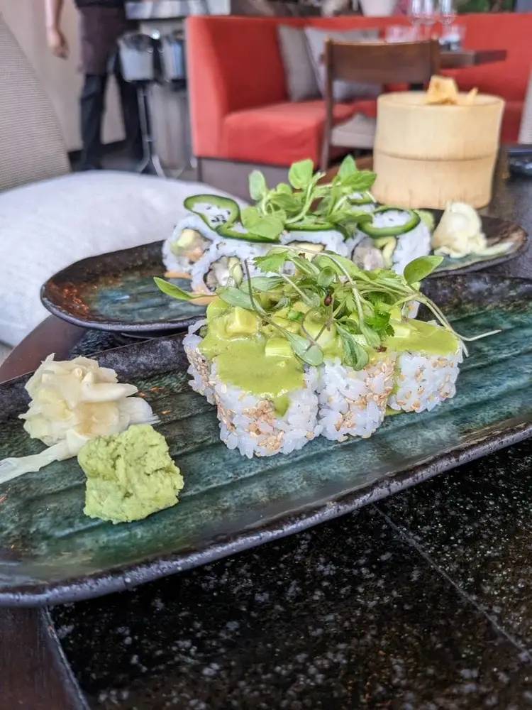 Sushi rolls topped with green sauce and microgreens sit on a dark ceramic plate with wasabi and ginger, displayed in a modern restaurant dining room