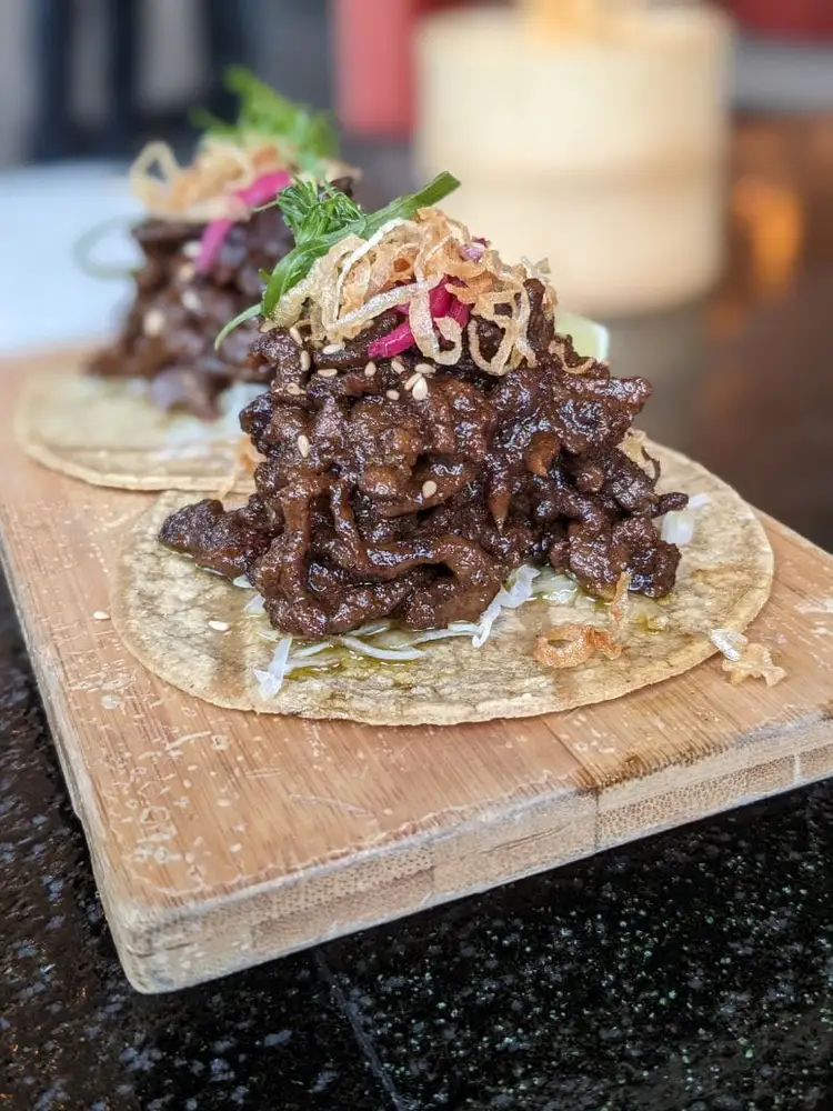 Shredded meat tacos topped with crispy noodles, pickled vegetables, and fresh herbs sit on a wooden board