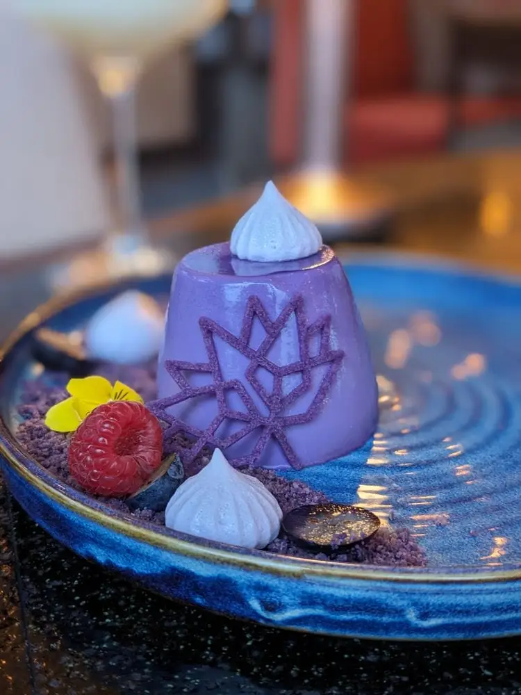 Purple pastry dome with piped white cream top sits on a blue plate garnished with a red raspberry, yellow flower, and decorative elements