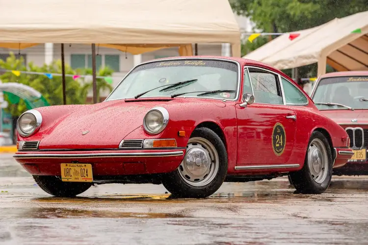 Red vintage sports car parked on wet pavement at an outdoor event with tents and other classic vehicles visible in the background