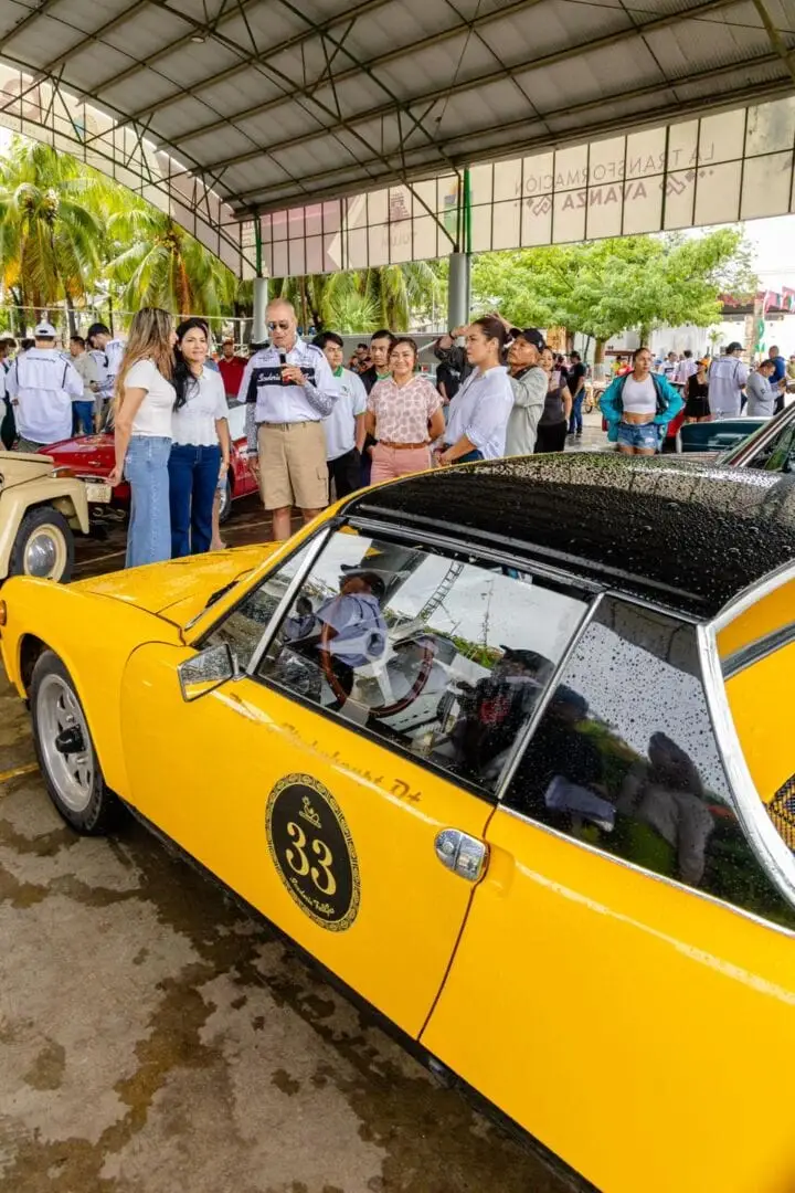 A yellow vintage car with an open hood is displayed under a metal-roofed structure while a crowd of people examines classic automobiles on display