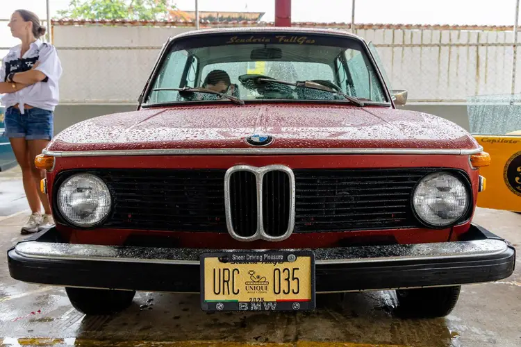A red BMW vintage car with distinctive dual circular grilles sits wet on pavement at an outdoor event, with spectators nearby