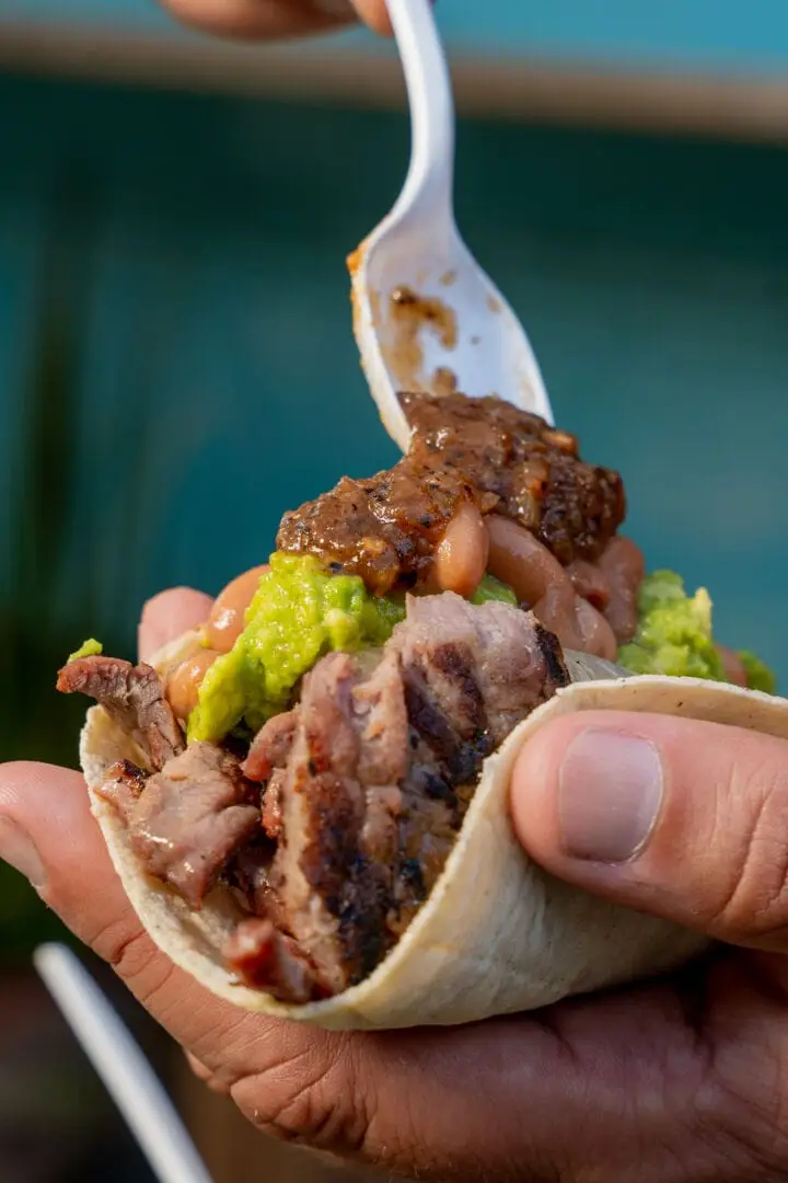 A hand holds a sandwich filled with sliced grilled meat and green sauce while another hand drizzles sauce from a white spoon above it A hand holds a sandwich filled with sliced grilled meat and green sauce while another hand drizzles sauce from a white spoon above it