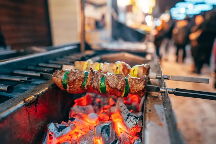 Meat and vegetable skewers cook over hot coals on a grill at an outdoor food event with people visible in the blurred background Meat and vegetable skewers cook over hot coals on a grill at an outdoor food event with people visible in the blurred background