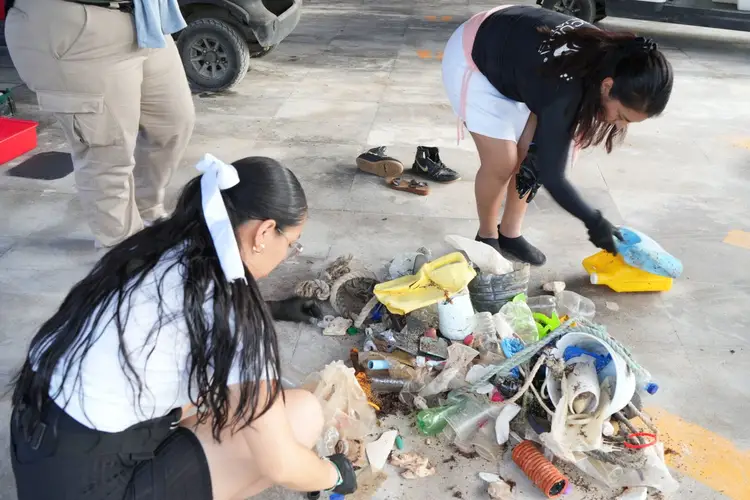 Two women collect plastic waste and debris from a concrete surface in a parking area, sorting items into piles with collection bags nearby