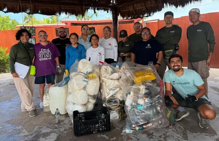 A group of volunteers stands behind bags and containers of collected marine debris and plastic waste under a thatched roof structure